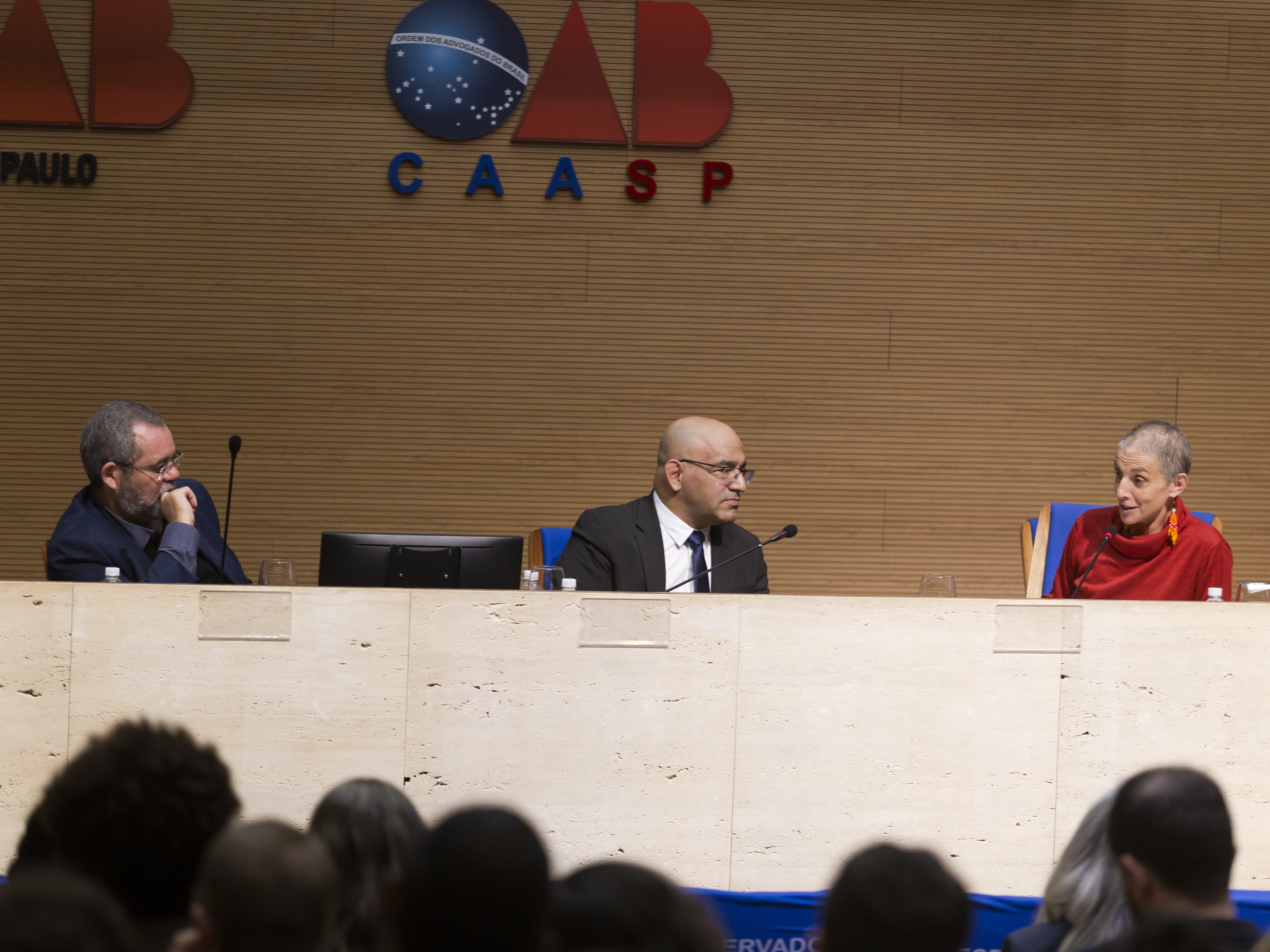 Grupo de três pessoas sentadas atrás de um balcão, a frente de uma parede com os símbolos "OAB São Paulo" e "OAB CAASP". Uma plateia assiste a fala das três pessoas.