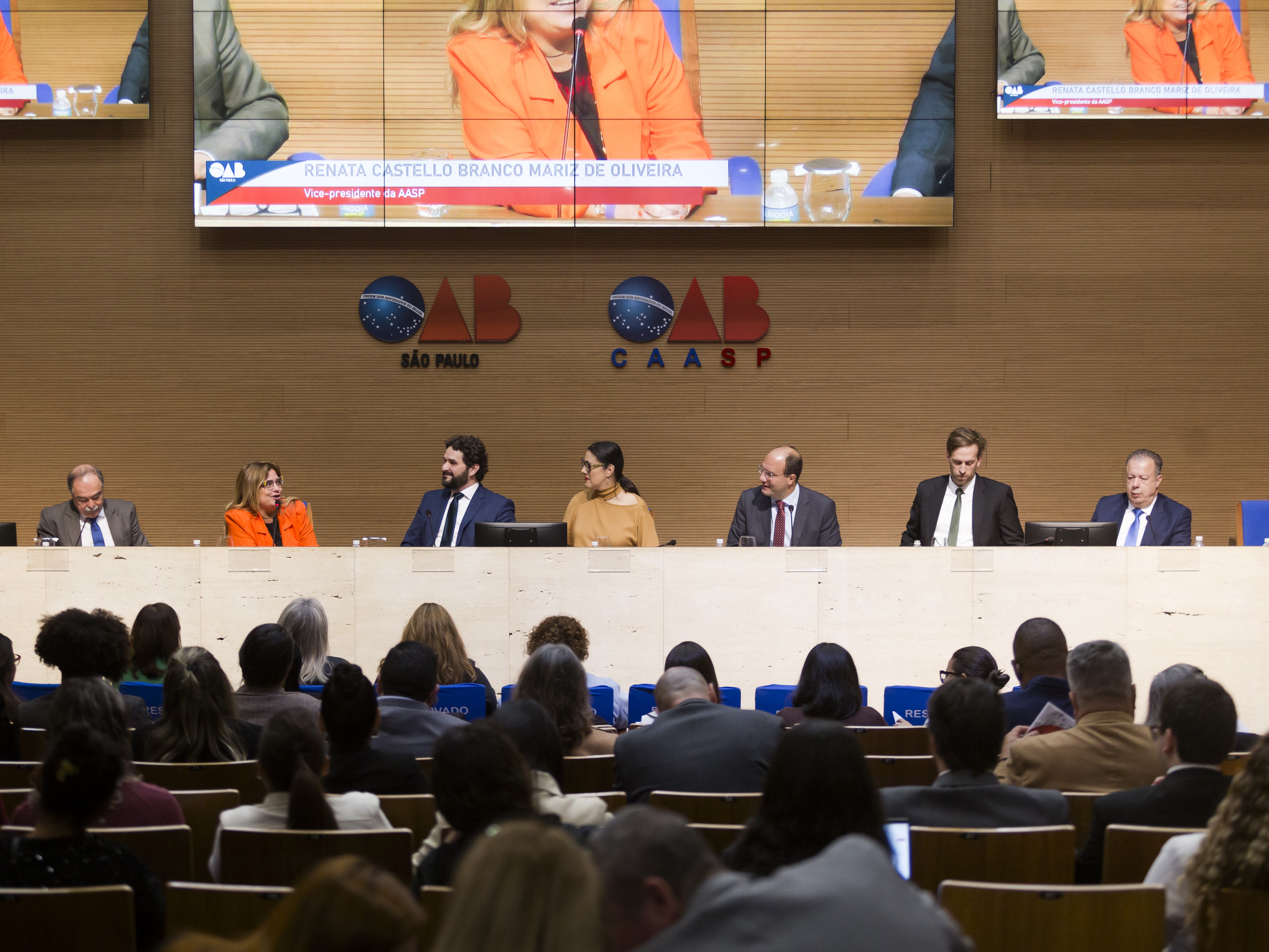 Grupo de sete pessoas sentadas atrás de um balcão, a frente de uma parede com os símbolos "OAB São Paulo" e "OAB CAASP". Uma plateia assiste a fala das sete pessoas.