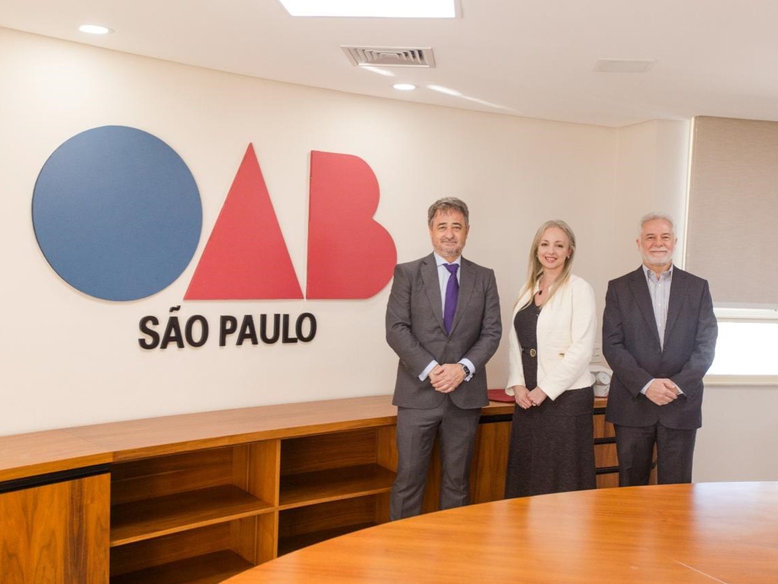 Três pessoas em roupas formais, posando atrás de uma mesa redonda com o símbolo "OAB São Paulo" na parede ao fundo.